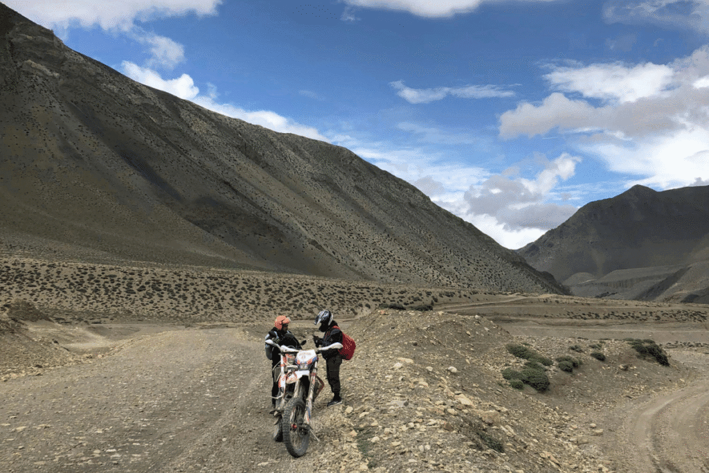 Two people with helmets are standing next to a red and white motorbike on a dirt road in a mountainous, arid landscape.