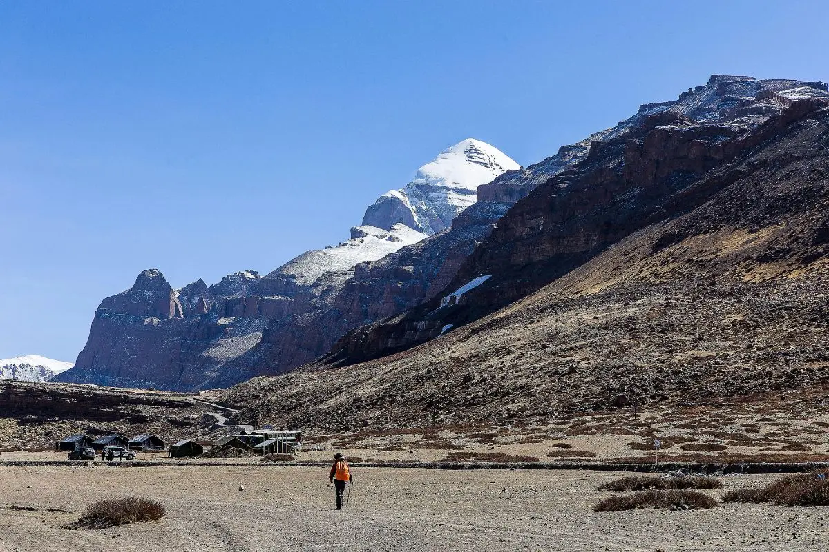 A lone trekker with an orange backpack walking toward the snow-capped peak of Mount Kailash under a clear blue sky.