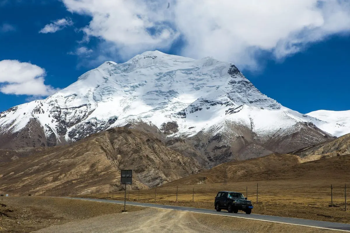 A black SUV driving on a paved road toward a massive, snow-covered Himalayan mountain range.